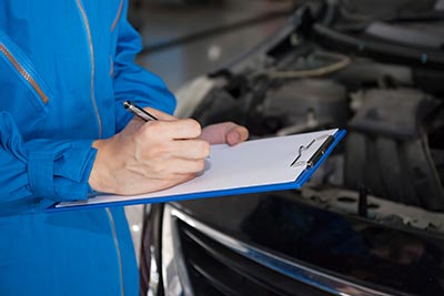 Carolina Collision and Frame Service | close up of man wearing a blue jumper writing on a tablet attached to a clipboard in front of a car's engine